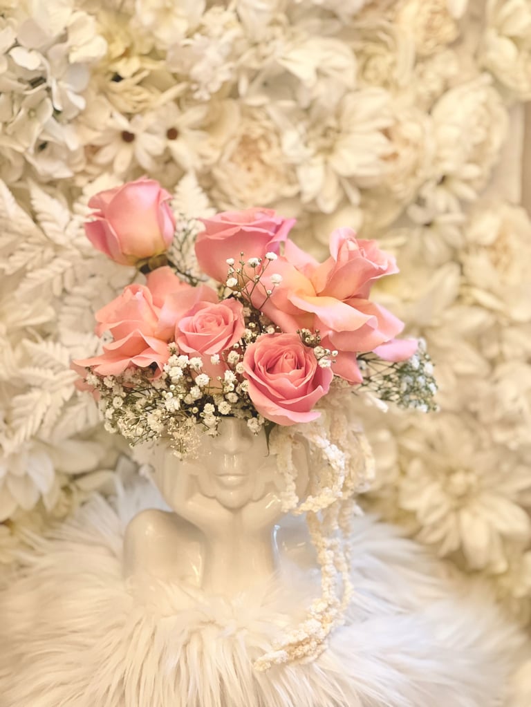 Pink roses and babys breath arranged in an ornate cream vase with pearls, displayed against white flowers