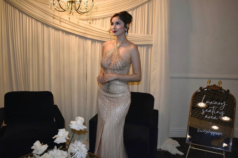 Woman in elegant beaded gray gown standing in luxurious room with white drapes, chandelier, and floral arrangements