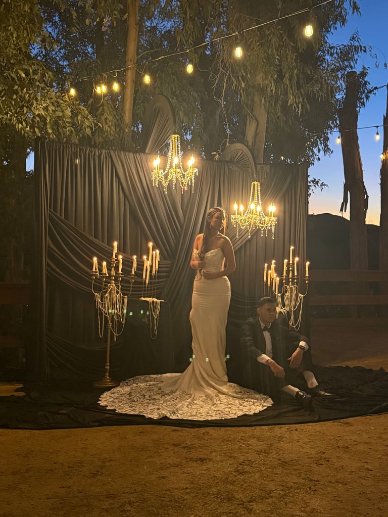 Bride in white gown and groom in dark suit pose under an ornate outdoor backdrop with chandeliers, draped fabric, and string lights at dusk