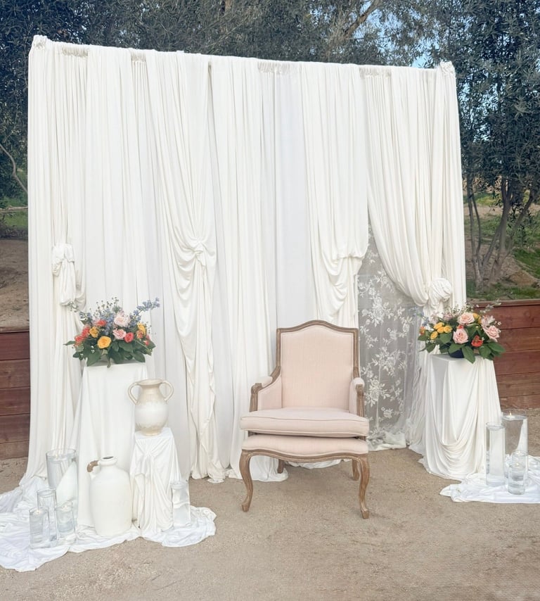Elegant wedding ceremony setup with white draped fabric enclosure, cream armchair, floral arrangements, and decorative vessel on ground.